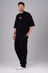 Black oversized t-shirt with short sleeves, featuring a rectangular red logo patch. Worn with black jeans and white sneakers.