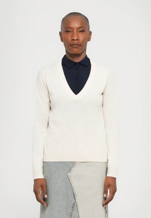 V-neck beige knit sweater with long sleeves, ribbed cuffs, and hem. Worn over a dark blue collared shirt, paired with a light denim skirt.