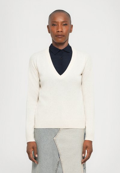 Pull en maille beige à col en V, à manches longues, avec des poignets et un ourlet côtelés. Porté par-dessus une chemise à col bleu foncé, associé à une jupe en denim clair.