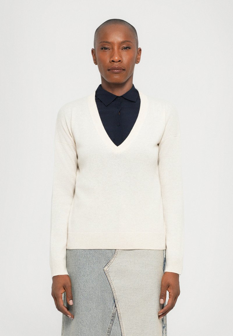 V-neck beige knit sweater with long sleeves, ribbed cuffs, and hem. Worn over a dark blue collared shirt, paired with a light denim skirt.