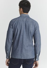 Mėlynas chambray marškinys, ilgomis rankovėmis, mygtukų rankogaliais, subtilia tekstūra, galinis vaizdas, pabrėžiantis tiesų siluetą ir yoko detales.