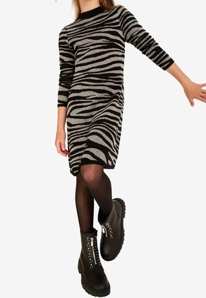 Fille portant une robe rayée zèbre noire et grise, des collants noirs et des bottines noires avec fermetures éclair à l'avant et détails cloutés.