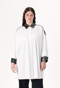 Hvit skjorte med en svart, teksturert krage og mansjetter. Har knapper foran og løs passform med lange ermer. Glatt stoff, minimalistisk design.