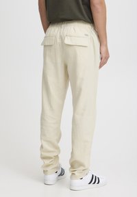 Lys beige joggebukser med avslappet passform, elastisk midje, to baklommer og myk, teksturert stoff. Brukt med hvite sneakers med svarte striper.