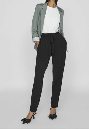 Mujer con blazer gris claro, camisa blanca, pantalones negros de talle alto con lazada frontal y tacones negros con lazo, de pie con las manos en los bolsillos.