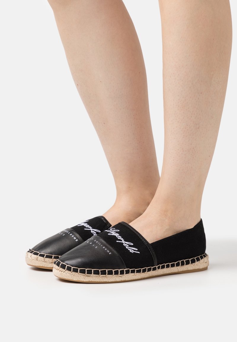 Jambes portant des espadrilles noires à enfiler avec des semelles blanches cousues et un texte blanc en écriture manuscrite à l'avant, debout sur une surface blanche.