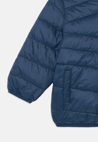 Veste matelassée bleu marine avec coutures en losange, dotée d'un poignet arrondi et d'une poche latérale. Texture lisse et brillante.