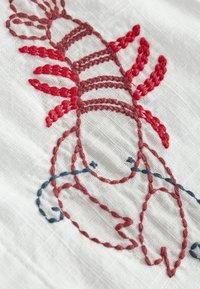 Broderie en fil rouge et bleu représentant une langouste sur un tissu blanc tissé, avec des points détaillés et une texture visible.
