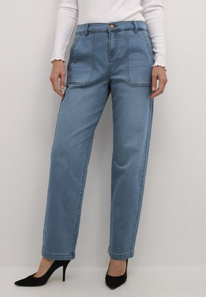 Vrouw in lichtblauwe rechte jeans met grote voorzakken, een witte lange mouwen top en zwarte hoge hakken.