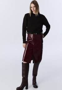 Un pull marron en peluche associé à une jupe crayon bordeaux brillante, arrivant au genou, avec une fente sur le devant, et des bottes à talons hautes assorties.