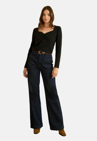 Fekete, hosszú ujjú felső négyzet alakú kivágással és ráncolt részletekkel, széles lábú sötét farmernadrággal és világosbarna magassarkú csizmával párosítva.