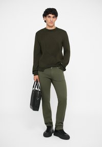 Sötétzöld, hosszú ujjú pulóver gyémántmintával, olíva zöld, slim fit nadrággal és fekete válltáskával párosítva. A modellen fekete sportcipők vannak.