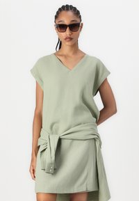 Abito verde chiaro con scollatura a V, maniche corte e dettaglio in vita annodato. Il tessuto sembra morbido e testurizzato, adatto per un abbigliamento casual.
