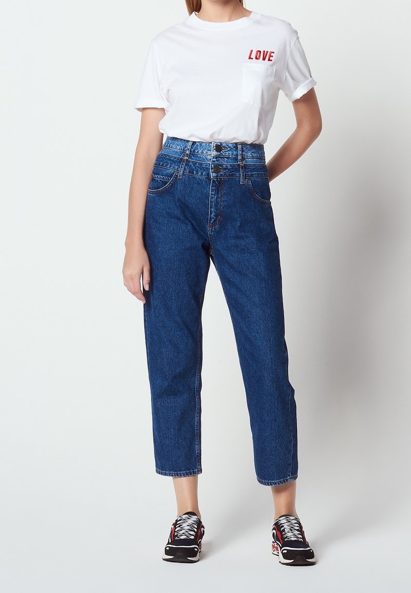 Personne portant un t-shirt blanc avec l'inscription "LOVE", un jean bleu taille haute avec boutons, et des baskets noires avec des touches rouges et blanches.