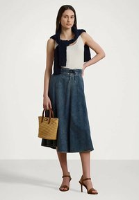 Sieviete ģērbusies baltā bezpiepšļotā topā, tumši zilā denima midi svārkos, jūras zila džemperī pār pleciem, brūnās sandales, tur rokassomu no austa materiāla.