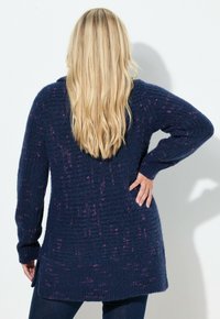 Długi granatowy sweter z dzianiny o teksturowanym wzorze, z delikatnymi różowymi plamkami i luźnym krojem. Bocznie wycięcia zwiększają swobodę ruchów.