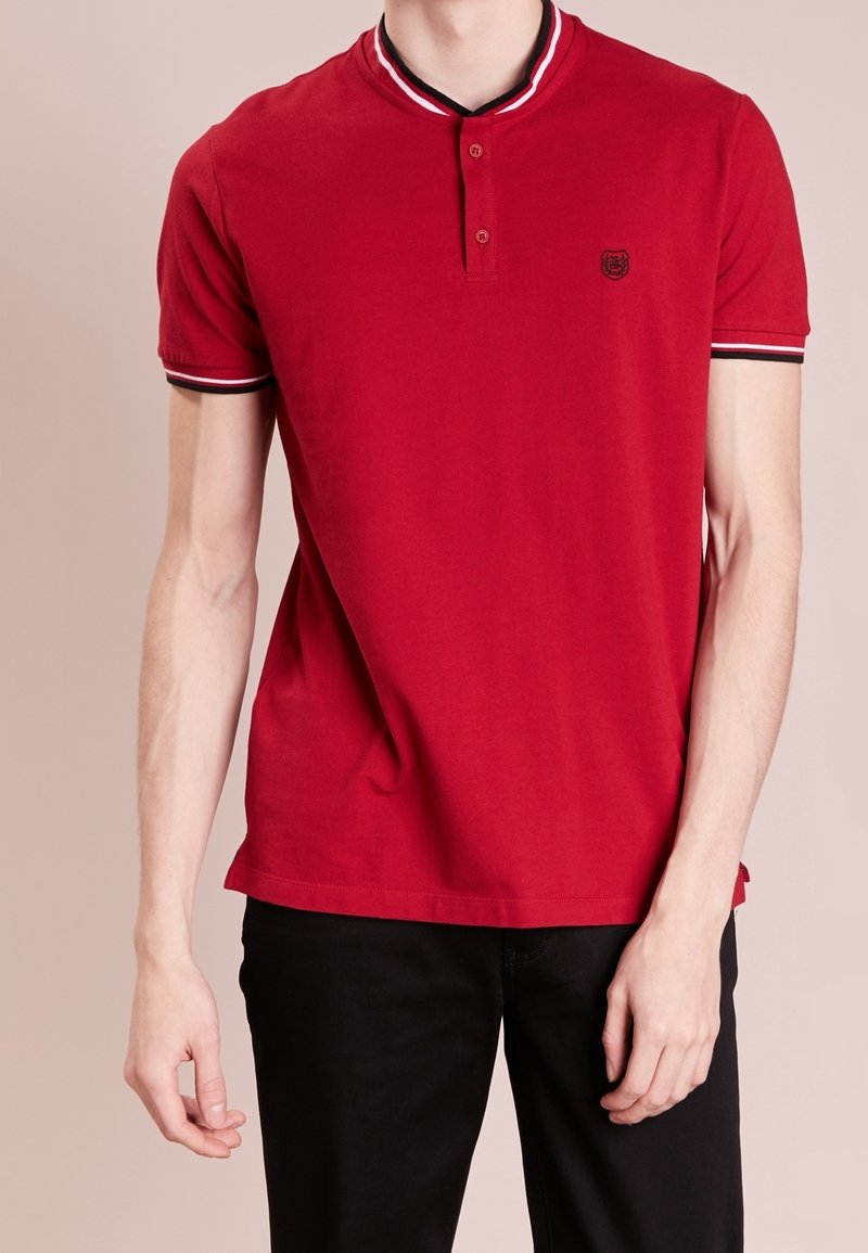 Röd bomulls-pikétröja med en tvåknapps-knäppning, kontrasterande svart och vit ribbad krage och ärmkanter, med en liten emblem på bröstet.