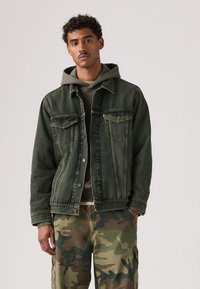 Σκούρο πράσινο τζιν μπουφάν με δύο τσέπες στο στήθος και μπροστινό κλείσιμο με κουμπιά, φορεμένο πάνω από γκρι φούτερ με κουκούλα, σε συνδυασμό με παντελόνια παραλλαγής.