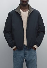 Navy bomber jacket with a light gray collar, zippered pockets, and ribbed cuffs, worn over a light beige sweater and blue jeans.