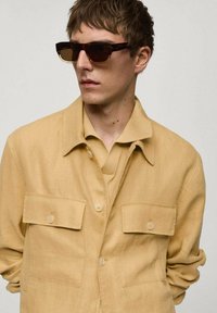 Svetle žlutá lněná košile s dvěma náprsními kapsami, nošená přes žebrovaný béžový top. Model má na sobě tmavé, hranaté sluneční brýle.