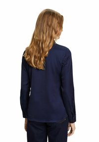 Femme aux longs cheveux blonds ondulés portant une chemise bleu marine à manches longues et un pantalon foncé, debout de dos sur un fond blanc.