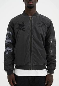 Black bomber jacket with embroidered eagle and text details, zip closure, ribbed cuffs, and a side pocket; smooth textile finish.
