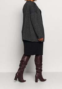 Cardigan grigio a maglia, oversize con spalle cadenti; abito nero aderente; stivali al ginocchio bordeaux lucidi con tacco a blocco e texture arricciata.