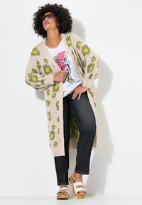 Beige neulecardigan, jossa on vihreitä leoparditäpliä, pitkät hihat, ylipitkä malli. Päällystetty valkoisen graafisen T-paidan ja tummien farkkujen kanssa, yhdistetty puu-kenkien kanssa.