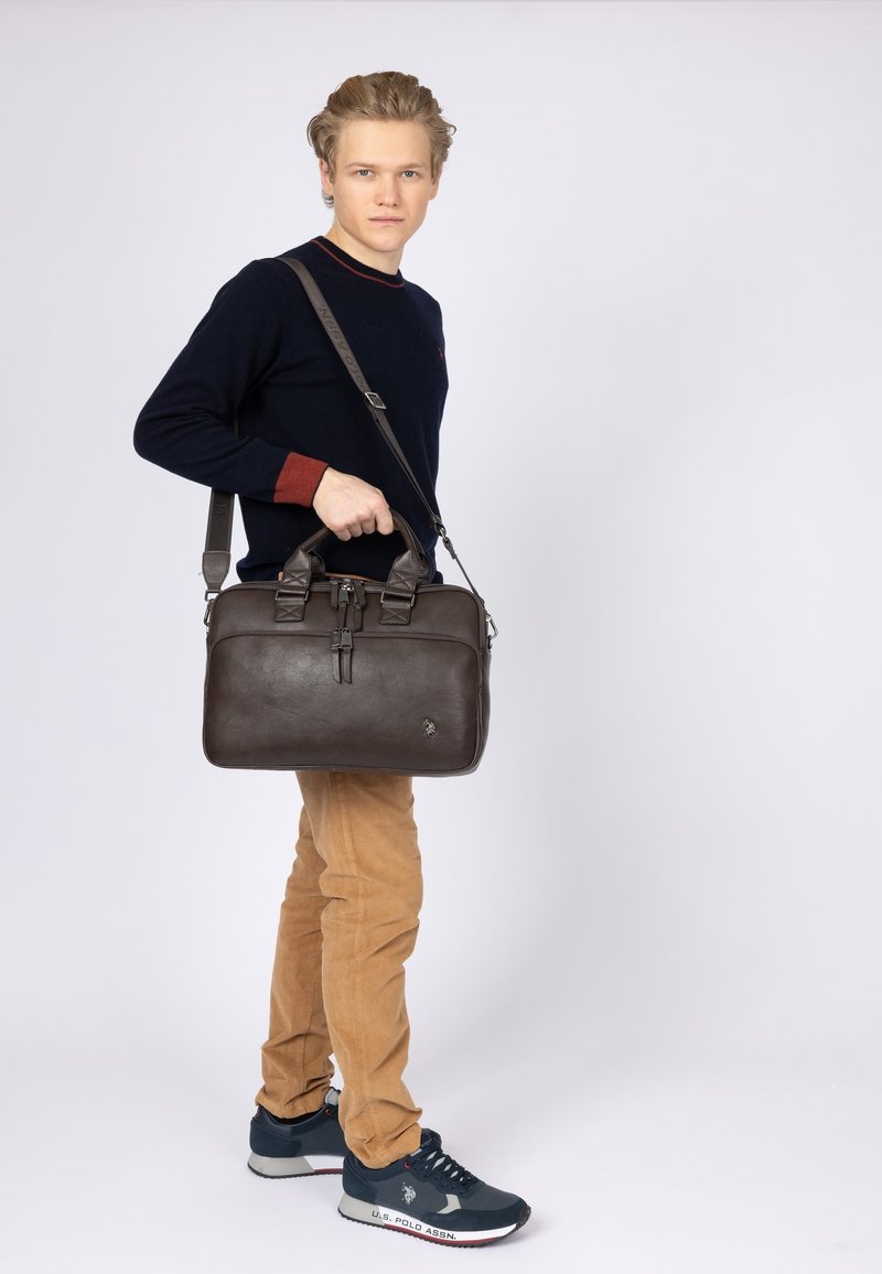 Borsa per laptop in pelle marrone con manici superiori doppi, tracolla removibile, chiusura con zip e dettagli cuciti, mostrata da un modello.