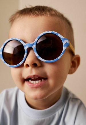 Malé dítě s velkými kulatými slunečními brýlemi s modro-bílým vzorem, mírně se usmívající, na neutrálním pozadí.