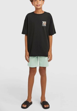 Črna oversized majica s majhnim grafičnim motivom na prsih, kombinirana s svetlo zelenimi kratkimi hlačami in črnimi natikači. Preprost in priložnosten videz.