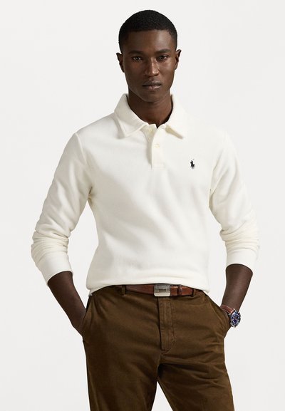 Crème polo shirt met een zachte textuur, lange mouwen, een knoopsluiting bij de kraag, en een klein zwart logo op de borst, gecombineerd met bruine broek.