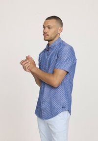 Chemise à manches courtes boutonnée en tissu bleu avec un petit motif blanc, présentant un col et des accents rouges subtils sur les boutons.