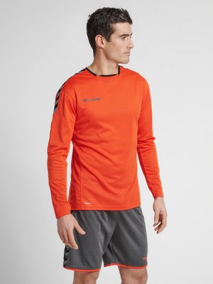 Mann trägt ein oranges Sportlangarmshirt und graue Shorts, steht und schaut seitlich vor einem schlichten Hintergrund.
