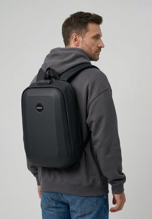 Mężczyzna w ciemnoszarej bluzie z kapturem i niebieskich dżinsach, niosący czarny plecak twardej konstrukcji VENEZA, stojący bokiem na jasnym tle.