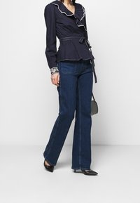 Marinblå peplumblus med rynkad halsringning och mönstrade ärmslut, kombinerad med utställda mörkblå jeans och svarta spetsiga klackskor.