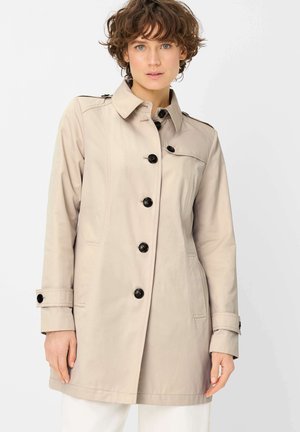 Frau mit kurzen lockigen Haaren, die einen beigen, knöpfbaren Trenchcoat mit Seitentaschen und schwarzen Knöpfen trägt, stehend vor einem weißen Hintergrund.