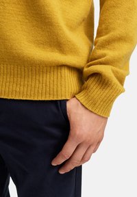 Żółty sweter z dzianiny z ściągaczami na mankietach i dolnym brzegu, noszony na ciemnogranatowych spodniach. Zbliżenie na teksturę materiału i dopasowanie wokół nadgarstka.