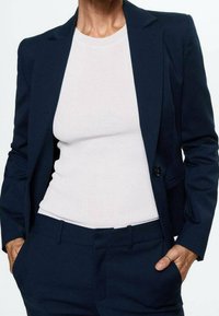 Blazer bleu marine et pantalon assorti avec un haut blanc côtelé. Le blazer présente une fermeture à un bouton et deux poches avant.
