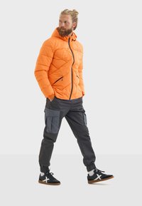 Chaqueta acolchada naranja con capucha y cremallera negra, combinada con pantalones cargo grises que tienen bolsillos laterales y zapatillas negras con detalles blancos.