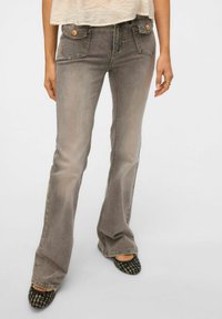 Grå denim flared jeans med to frontlommer, stikninger og bronze knapdetaljer, parret med teksturerede sorte flade sko.
