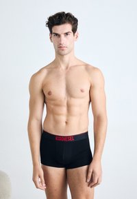 Schwarze Boxerbriefs mit rotem Bund, auf dem das Markenlogo zu sehen ist. Der Stoff sieht glatt und enganliegend aus, mit einem figurbetonten Design.