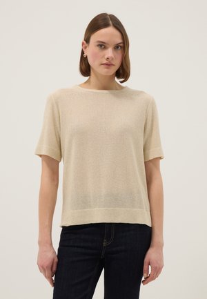 Mujer con cabello corto y castaño lleva un top beige de punto de manga corta y vaqueros oscuros, de pie frente a un fondo neutro.