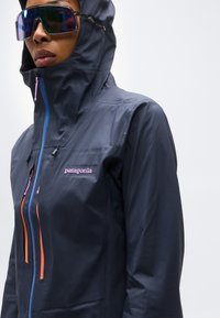 Άτομο που φοράει σκούρο μπουφάν Patagonia με κουκούλα, με πολύχρωμες λεπτομέρειες φερμουάρ και μεγάλα ανακλαστικά γυαλιά ηλίου, μπροστά σε απλό φωτεινό φόντο.