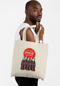 Beżowa torba z bawełny z kolorową grafiką butelek Coca-Coli oraz okr�ągłym logo z napisem "Lodowata Coca-Cola na sprzedaż".