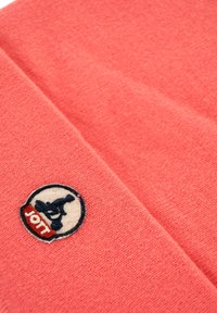 Primo piano di un tessuto a maglia corallo con una piccola toppa ricamata rotonda che presenta uno sciatore alpino nero e il testo rosso "JOTT".