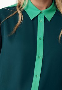Dark green blouse with a contrasting bright green collar and placket. Features buttons along the front and a smooth, soft fabric texture.