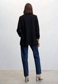 Blazer lungo nero con vestibilità rilassata, abbinato a jeans blu a vita alta e stivaletti beige con tacco, visto da dietro.