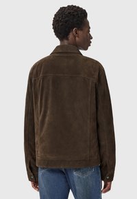 Mand med kort krøllet hår iført en mørkebrun ruskindsjakke og blå jeans, som vender væk mod en ensfarvet lys baggrund.