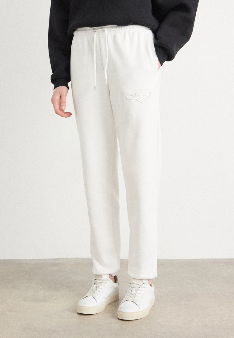Pantaloni della tuta bianchi con cintura regolabile, decorati con un logo ricamato. Abbinati a sneakers bianche su uno sfondo chiaro e neutro.